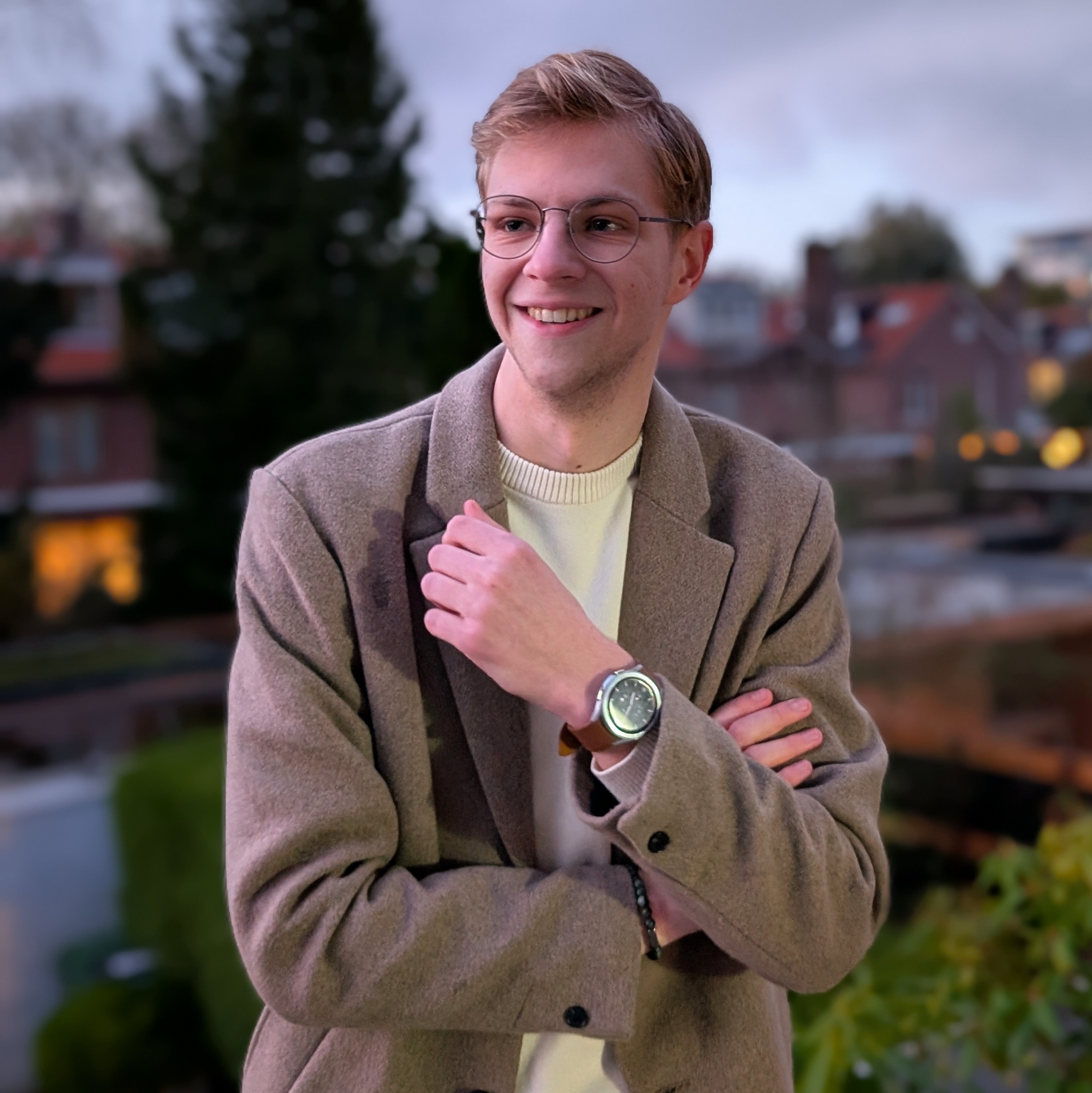 Een portret van Bence die met zijn armen over elkaar lachend naar rechts kijkt. Op de achtergrond is een wazige stedelijke omgeving te zien met gebouwen en groen. De foto is genomen bij schemering.