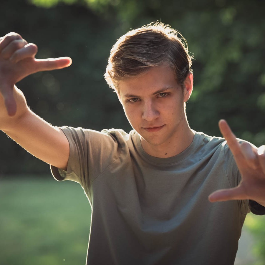 Een close-up portret van Bence die in de camera kijkt en met zijn handen een kader vormt. Hij staat buiten in een natuurlijke omgeving.