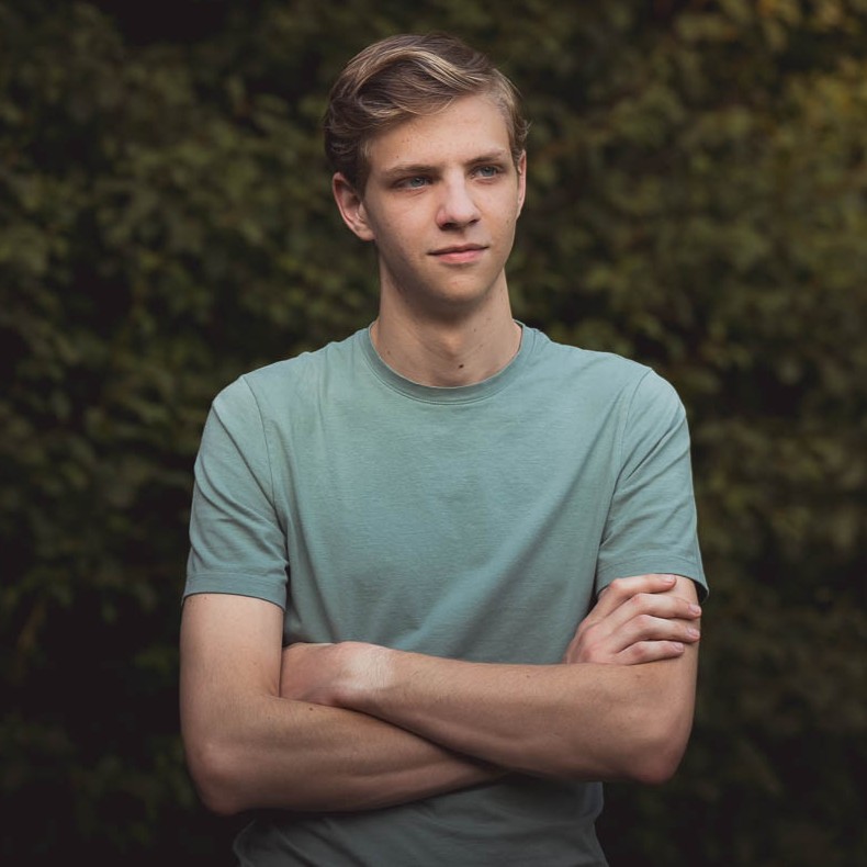 Een halflang portret van Bence die met gekruiste armen naar links kijkt. Hij staat buiten in een natuurlijke omgeving.